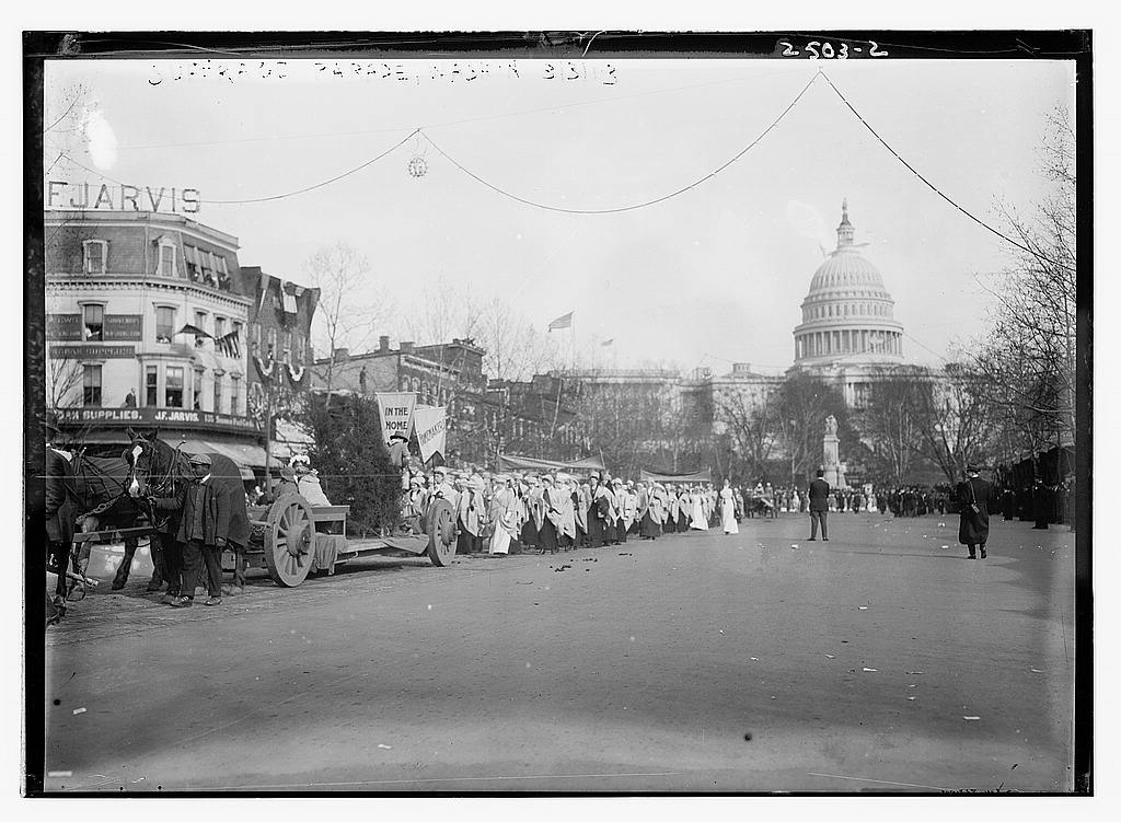 Women walking in a suffrage parade with banners and a crowd watching from the street.